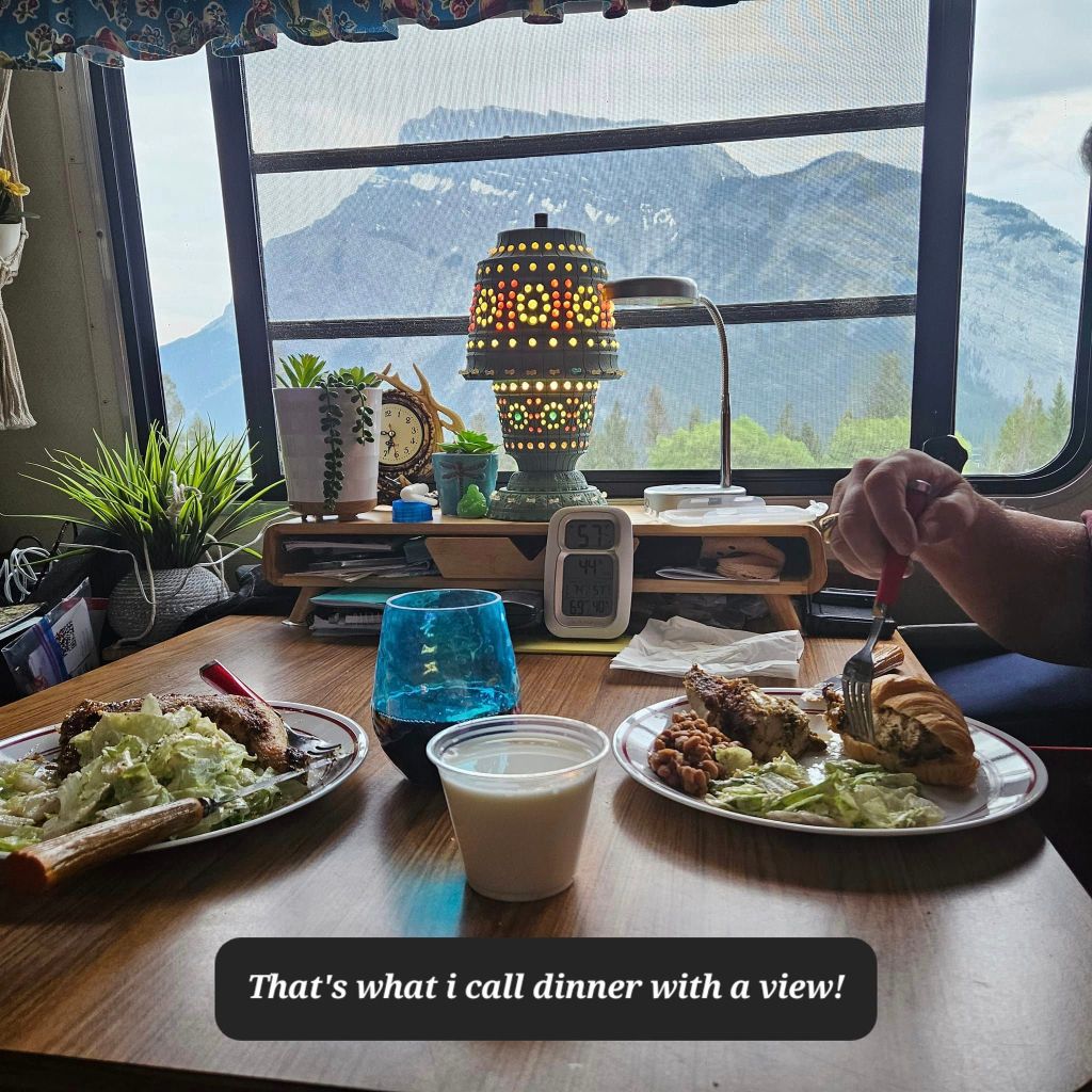 A cozy dining setup with two plates of food on a wooden table, featuring a scenic mountain view through a window. The table includes a colorful lamp, plants, and drinks.