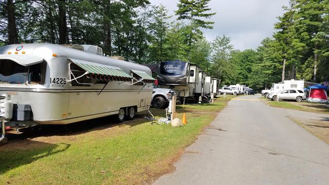 A row of recreational vehicles and trailers parked along a grassy campsite road, surrounded by trees and a cloudy sky.