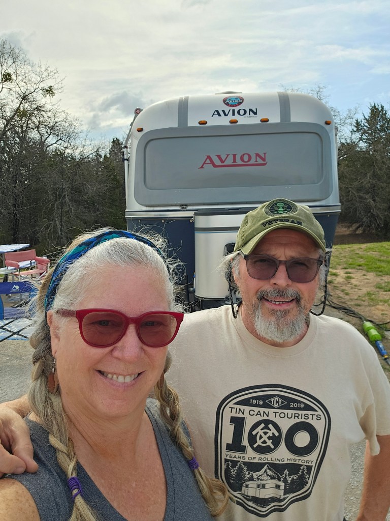 A couple poses for a selfie in front of a vintage Avion travel trailer, with trees and a cloudy sky in the background.