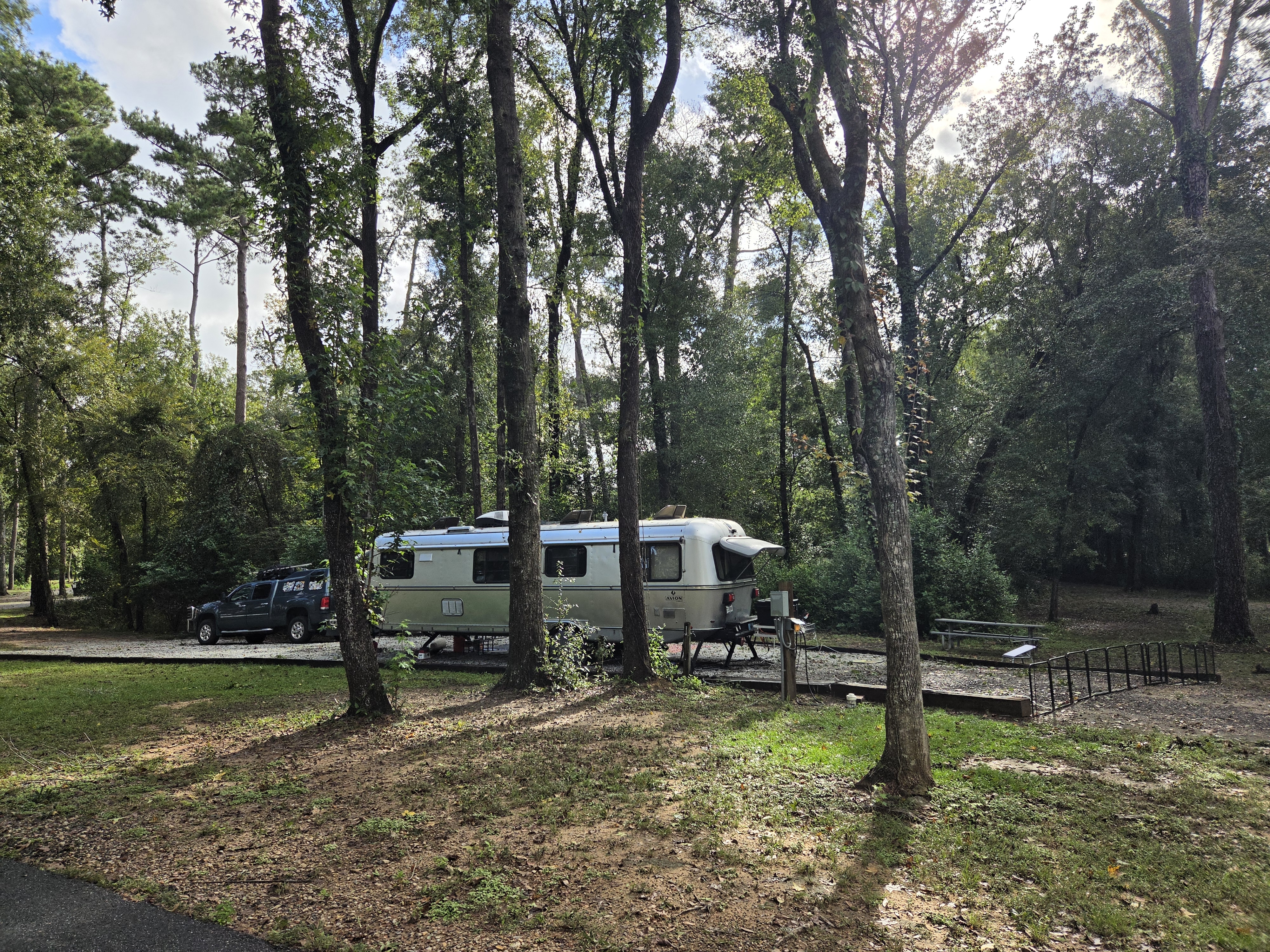 A silver travel trailer parked on a gravel pad amidst tall trees in a campground, with a black pickup truck nearby.